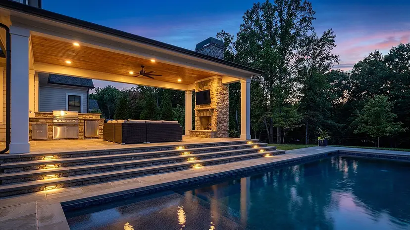 Covered patio with a cedar ceiling and stone fireplace above lit steps leading down to a pool at twilight